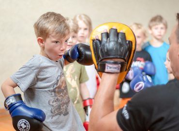Kind in blauen Boxhandschuhen schlägt auf die Pratzen des Trainers